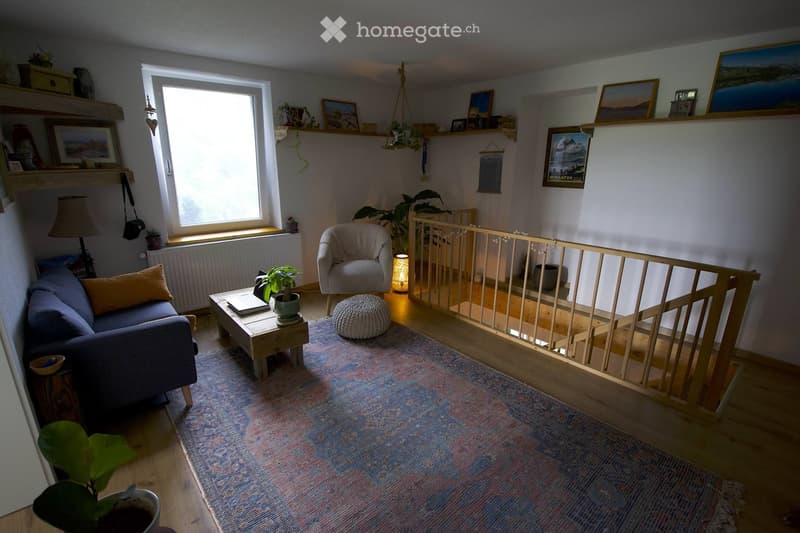 Cozy living room with a blue sofa, wooden coffee table, armchair, ottoman, and a small table lamp. Shelves with picture frames and plants are visible, along with a colorful rug and a wooden baby gate.