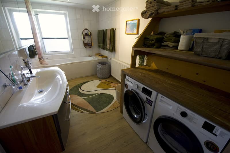 Bathroom with a sink, bathtub, washing machine, dryer, shelves with towels, and a window with blinds.