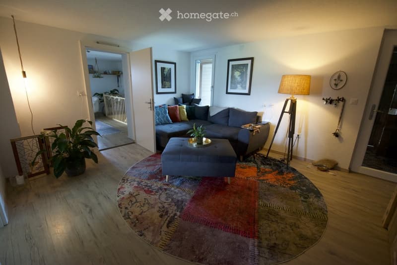 Living room with a grey sectional sofa, blue ottoman, floor lamp, small table with a plant, and picture frames on the wall.