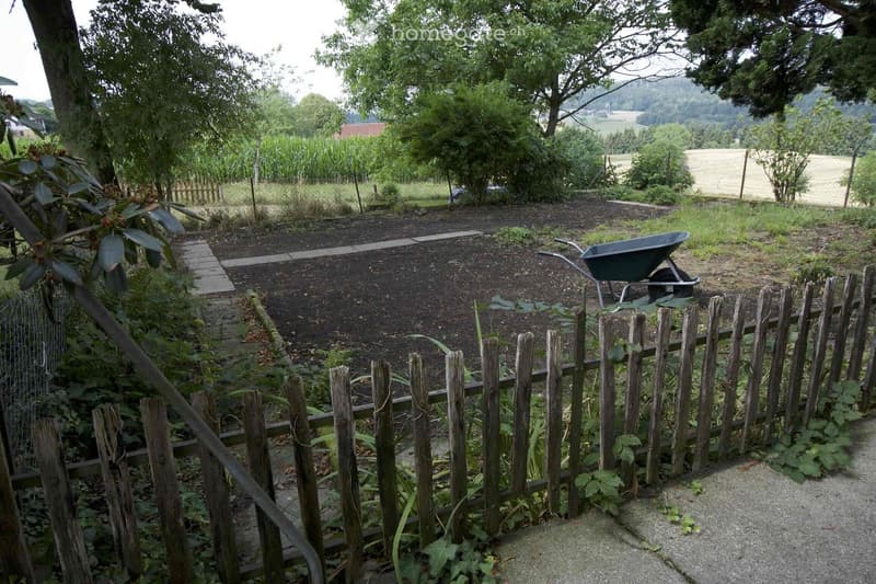 Garden area enclosed by a wooden fence, featuring a wheelbarrow, garden bed, and surrounding greenery with trees and open fields in the background.