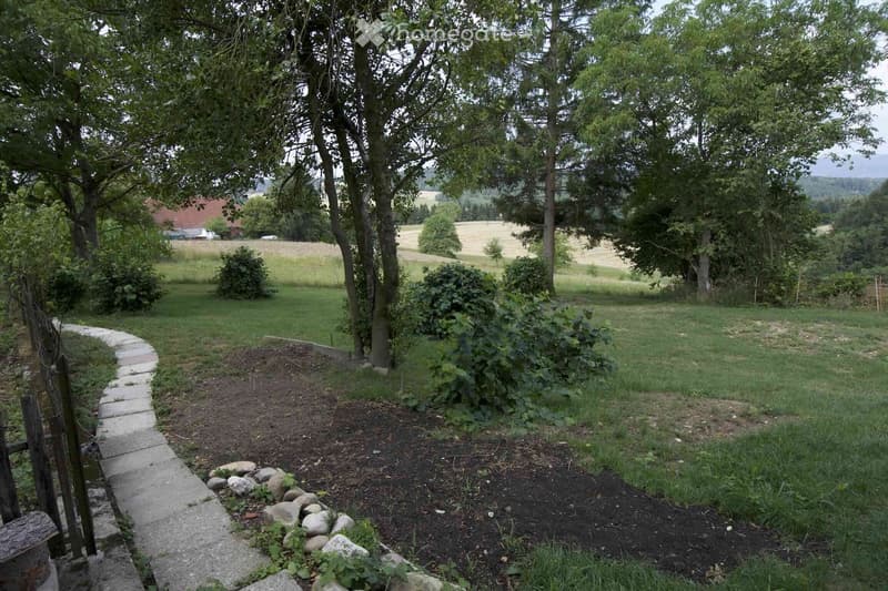 A garden area with a stone pathway, trees, bushes, and a grassy lawn, surrounded by a rural landscape with fields and distant hills.