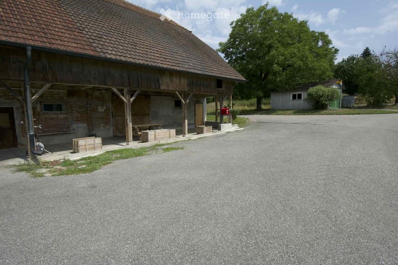 A rustic wooden barn with a tiled roof, brick walls, and a covered porch area. The building features wooden beams, a wooden door, and a small window. There is a gravel driveway leading to the barn, with a small wooden bench and crates near the entrance. T