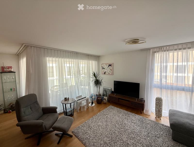 Modern living room with large windows, grey sofa, armchair, coffee table, piano, fan, TV, cabinet, and a rug.