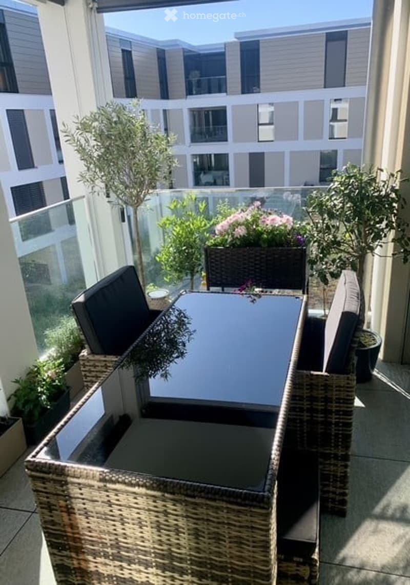 Balcony with wicker furniture, glass railing, potted plants, and a view of neighboring buildings.
