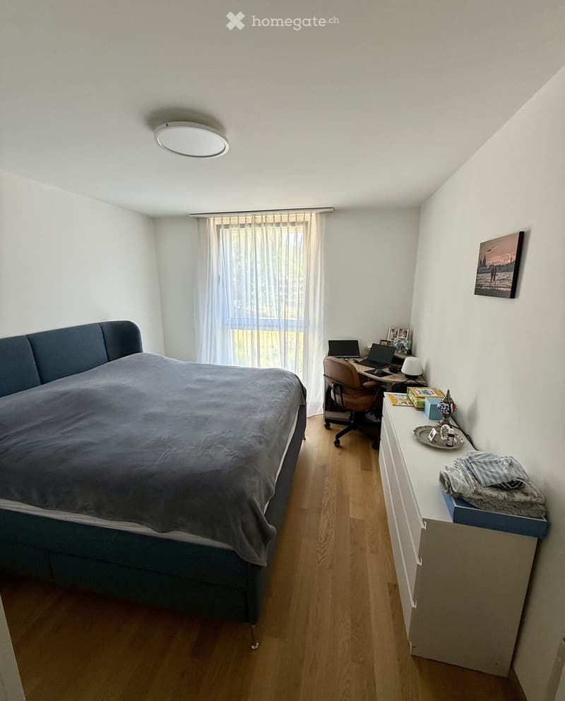 Cozy bedroom with a large bed, wooden floor, desk with laptop, chair, nightstand, and decorative items.