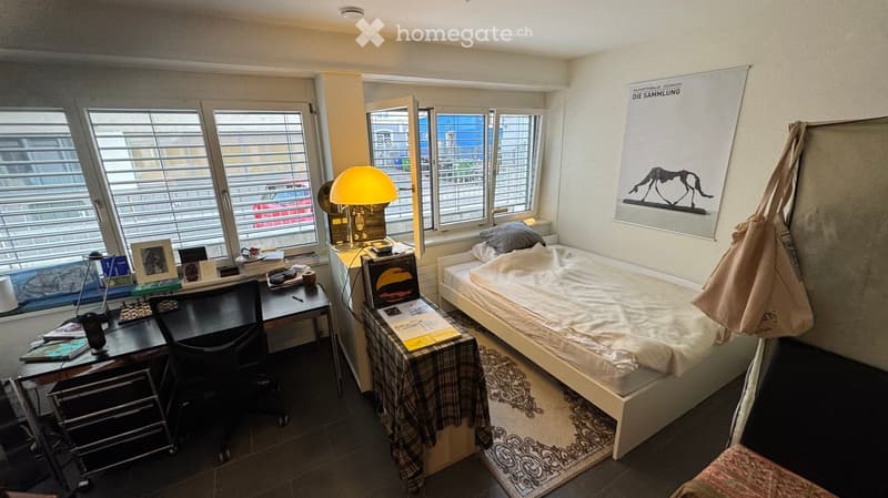 Cozy bedroom with a single bed, desk, chair, lamp, and various personal items. Large windows with blinds provide natural light.