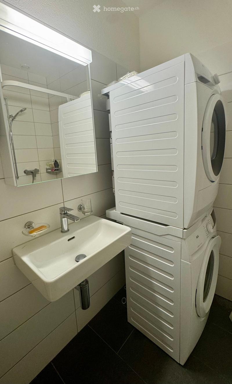 Modern bathroom with a white sink, faucet, mirror, and stacked white washing machine and dryer.