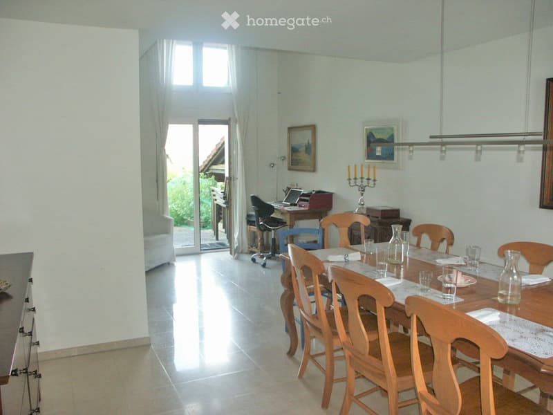 A well-lit dining room with a long wooden dining table surrounded by wooden chairs, a laptop on a desk, and a candle holder on a side table. The room features large windows with white curtains and a view of greenery outside.