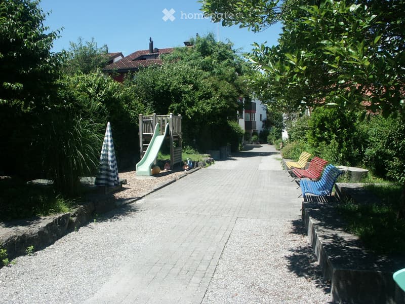 A well-maintained garden area with a paved pathway, a small playground with a slide, outdoor seating including chairs and tables, and lush greenery surrounding the area.