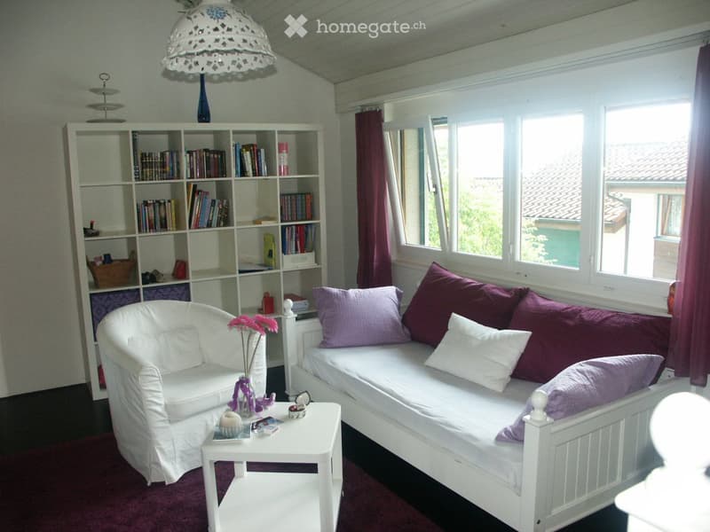 Cozy living room with a white sofa bed, a white armchair, a small table, and a bookshelf filled with books. The room features large windows with purple curtains and a decorative hanging lamp.
