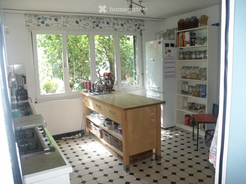 Bright kitchen with a wooden island, white refrigerator, blender, mixer, and shelves filled with jars and books. The room features a black and white checkered floor and large windows with blue and white patterned curtains.