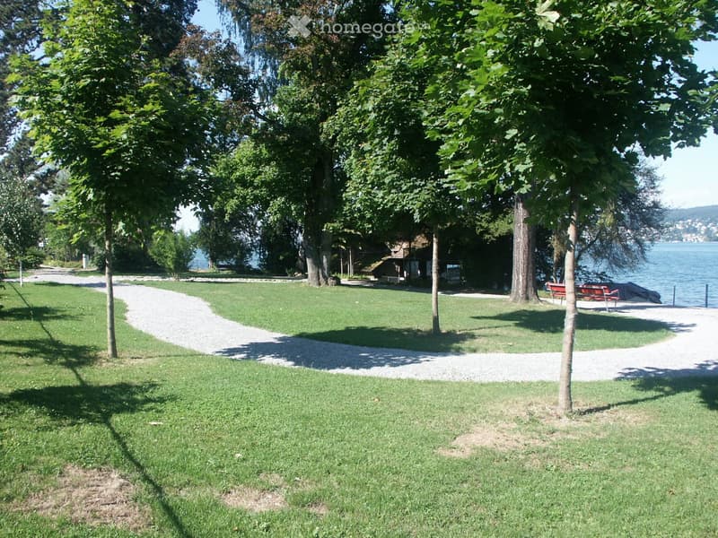 A well-maintained park area with green grass, trees, a gravel pathway, and a view of a body of water in the background.