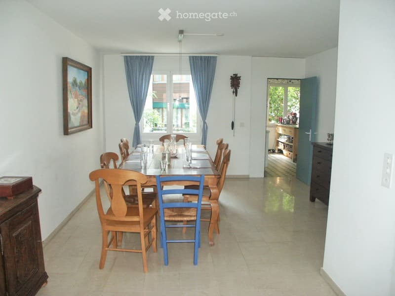 Dining room with a long wooden table set with glassware and chairs, large windows with blue curtains, and a view of greenery outside.