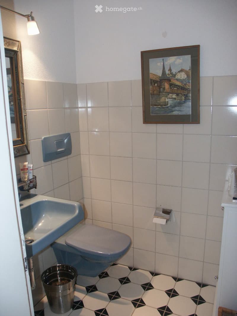 A small bathroom with white tiled walls, a sink, a toilet, a mirror, a framed picture, a waste bin, and a light fixture.