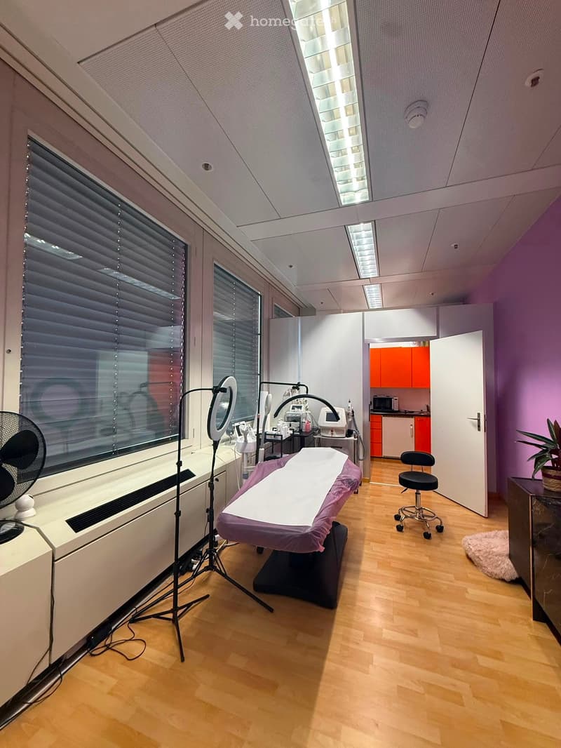 A well-lit, modern treatment room with a purple treatment table, a black stool, a ring light on a tripod, a fan, a small cabinet, a microwave, and a potted plant. The room features large windows with blinds and a sleek, contemporary design.