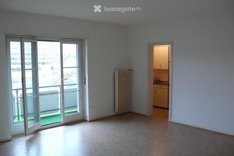 Empty living space with large sliding glass doors leading to a balcony, a radiator, and an adjacent small kitchen area.