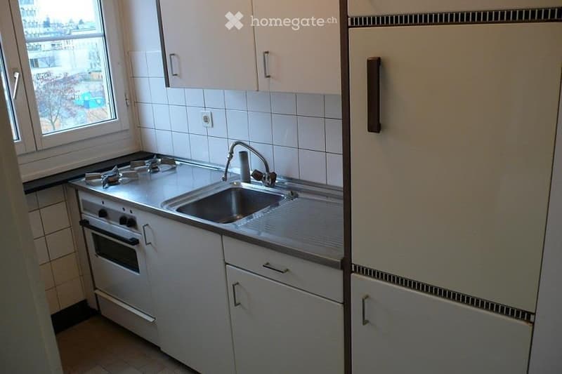 Compact kitchen with white cabinets, a gas stove, a sink, and a refrigerator. The space features tiled walls and a window offering a view of the outside.