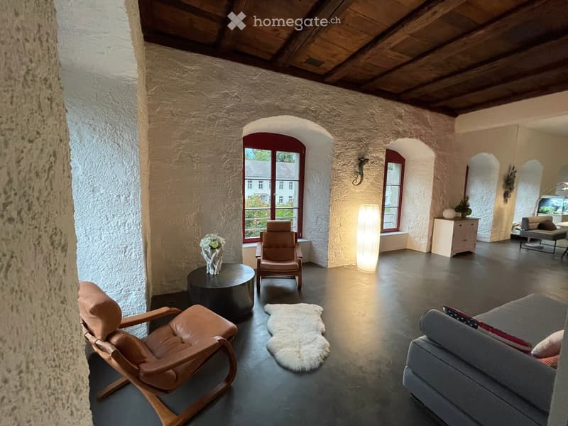 Living room with exposed wooden beams, stone walls, large windows, leather chairs, sofa, coffee table, floor lamp, and decorative rug.