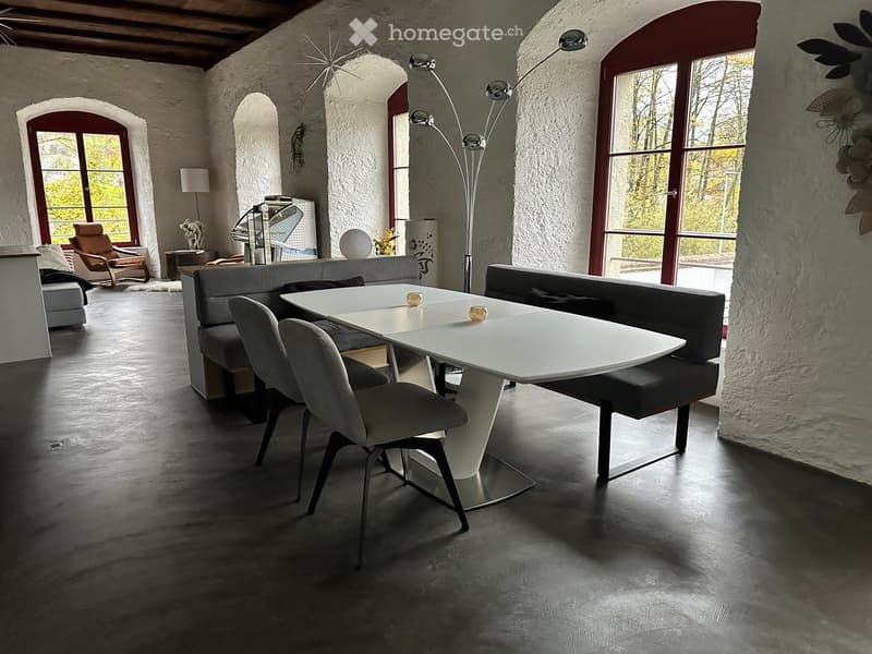 Modern dining area with a white table, grey chairs, and a grey sofa, featuring large windows and a sleek floor lamp.