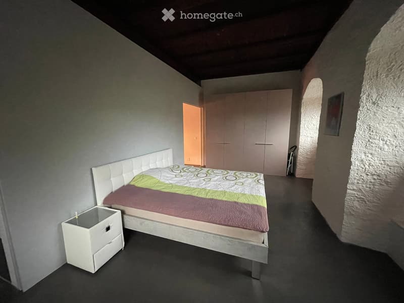 Modern bedroom with a white upholstered headboard bed, green and purple bedding, a white nightstand, and a large wardrobe. The room features dark flooring and exposed stone walls.