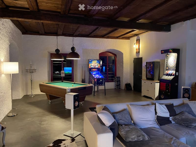 A spacious, modern living room with a white sectional sofa, a coffee table, a pool table, arcade machines, and a dartboard. The room features exposed wooden beams, pendant lights, and a cozy ambiance.
