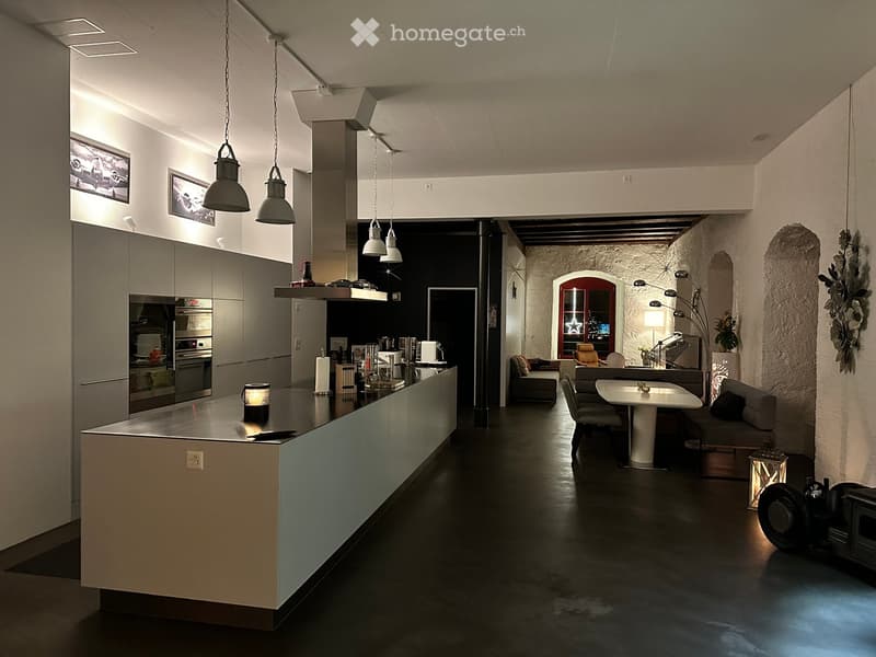 Modern open-plan kitchen and living area with a central island, pendant lights, a sofa, a dining table, and chairs. The kitchen features stainless steel appliances, including an oven, microwave, and fridge, with sleek cabinets. The living area has a cozy