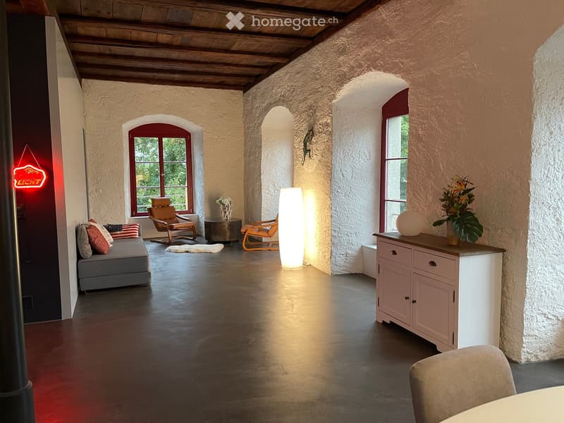 A spacious, well-lit room with exposed wooden beams, stone walls, and large arched windows. Features include a grey sofa with colorful pillows, wooden chairs, a small round table, a tall floor lamp, and a white cabinet with a vase of flowers.