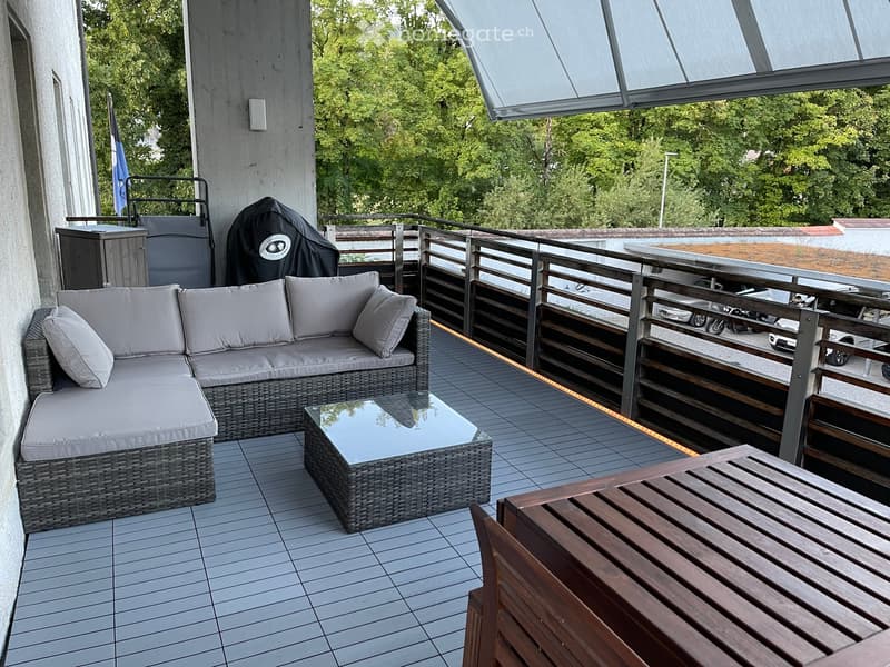 Balcony with a grey wicker sofa, glass coffee table, and wooden table, surrounded by a metal railing and lush greenery.