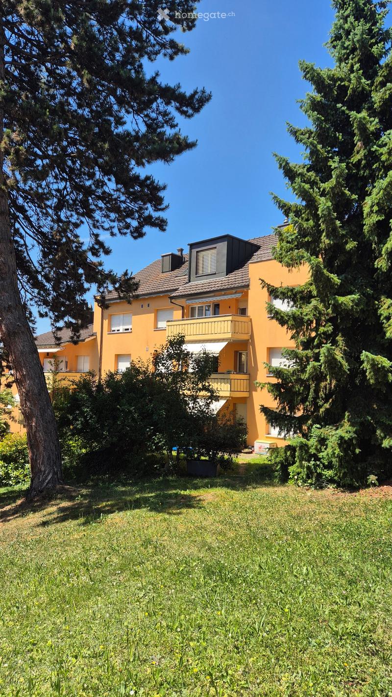 Two-story yellow house with a brown roof, multiple windows, and a balcony on the upper floor, surrounded by trees and greenery.