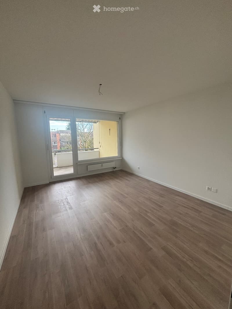 Empty room with wooden flooring and beige walls, featuring a window with a view of the outside.