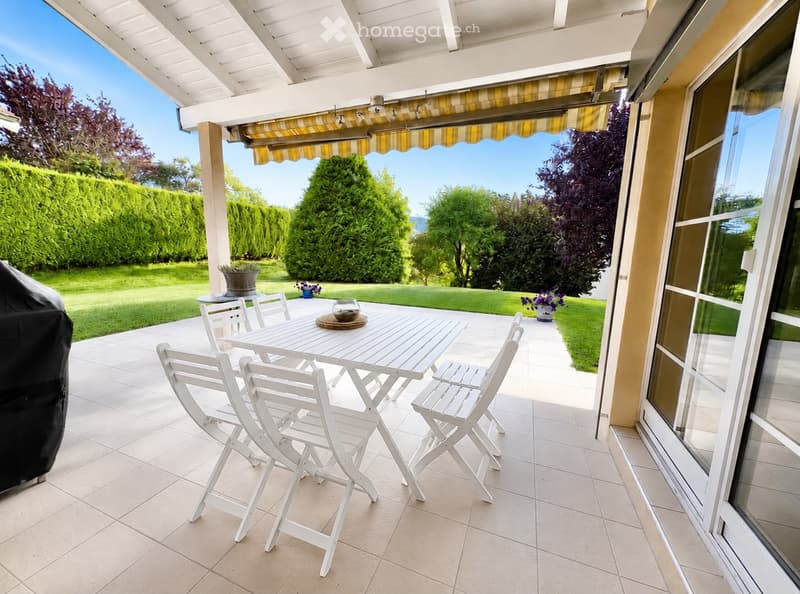 A covered patio with a white table and chairs, surrounded by a well-maintained garden and trees.