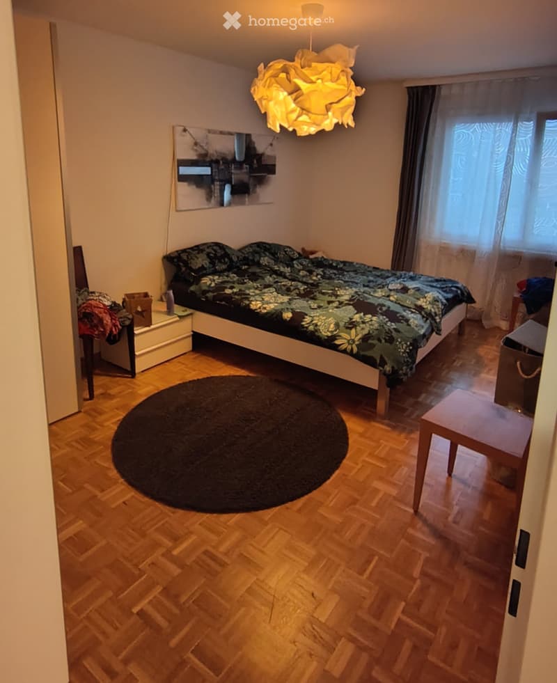 Cozy bedroom with wooden flooring, a large bed with floral bedding, a nightstand, a round rug, a chair, and a small table near the window.