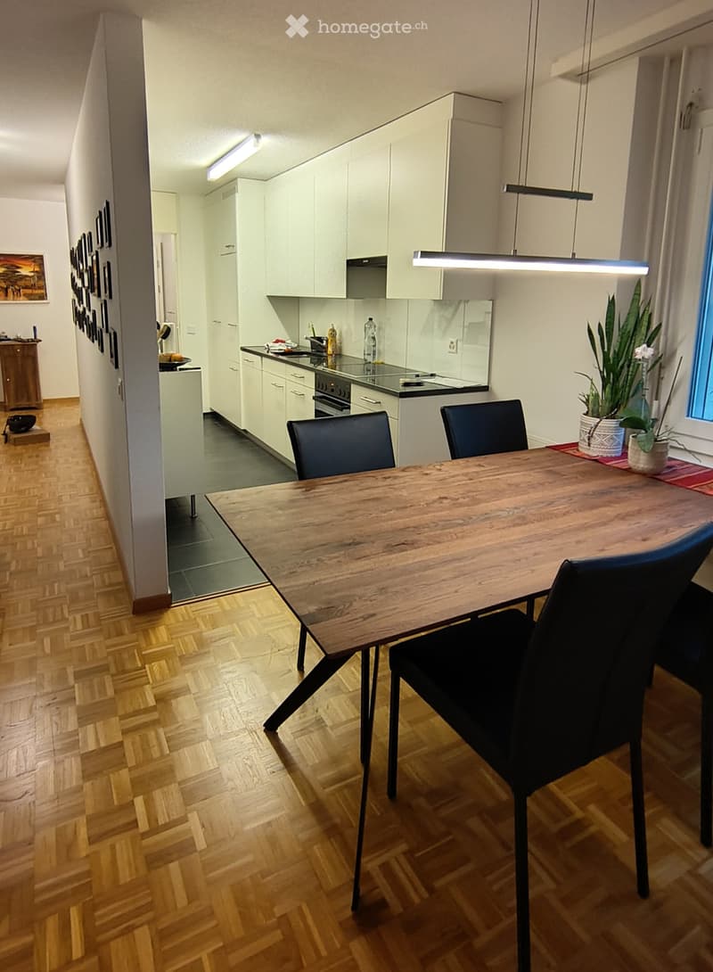 Modern kitchen and dining area with light-colored cabinets, a wooden dining table, black chairs, and a potted plant near the window.
