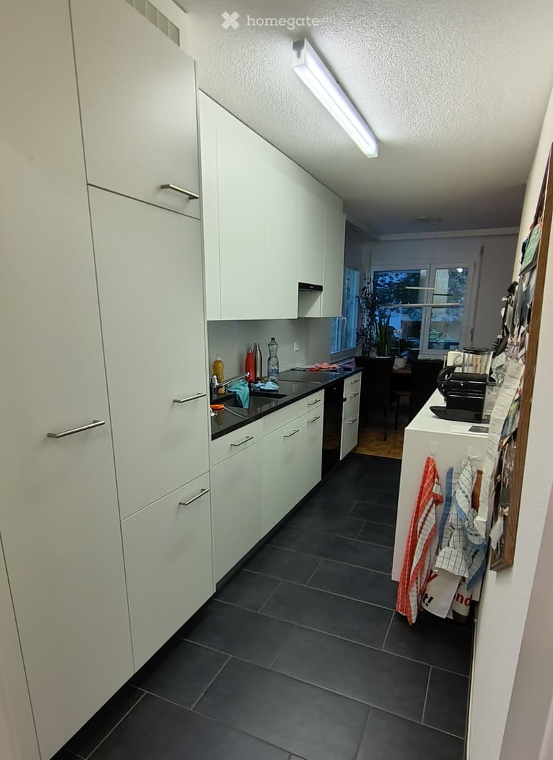 Modern kitchen with white cabinets, dark tiled floor, and stainless steel appliances. Visible items include a sink, faucet, coffee maker, and a small dining area with a table and chairs.