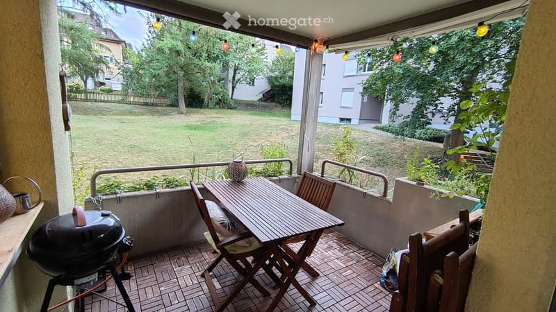 Balcony with wooden table, chairs, grill, and string lights, overlooking a green lawn and neighboring houses.