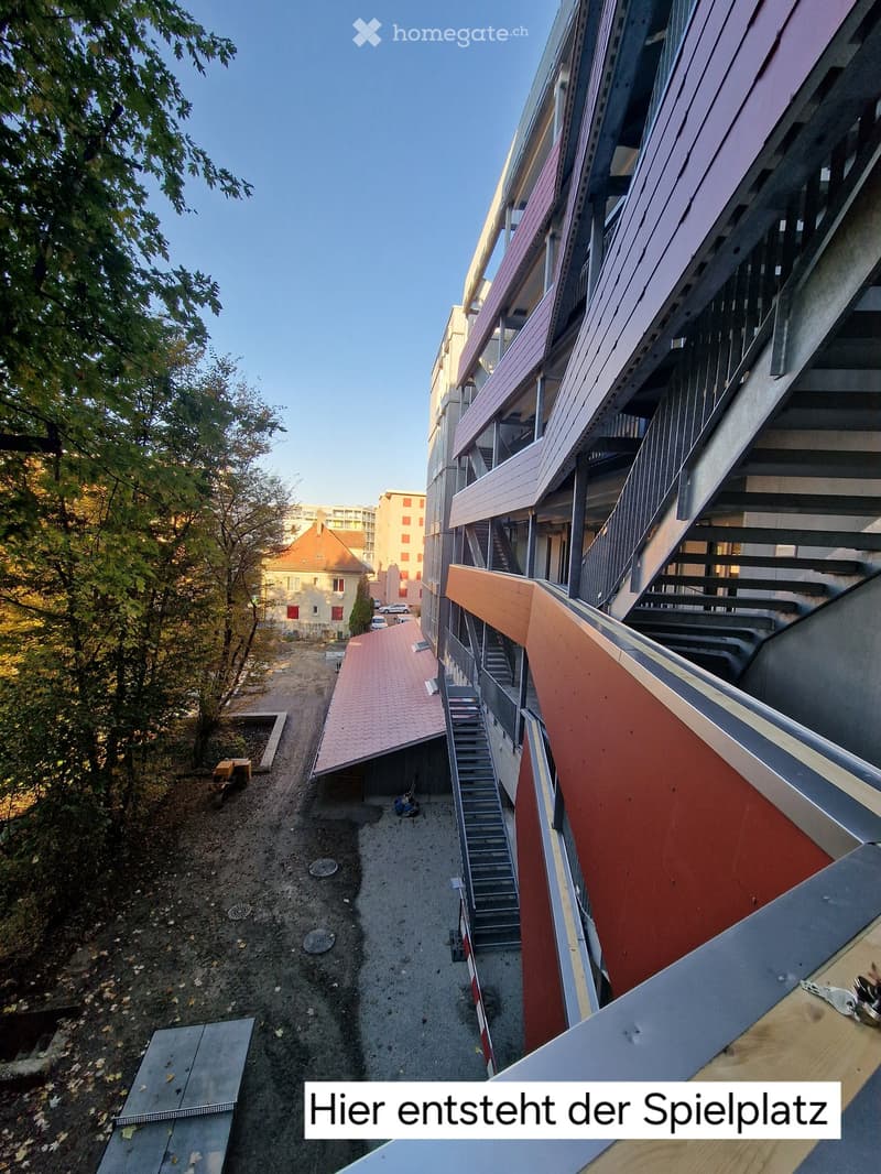 Modern multi-story building with a staircase leading to a rooftop area, surrounded by trees and a partially constructed playground area.