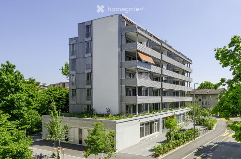 Modern multi-story residential building with balconies, large windows, and a raised ground floor with greenery and parking area.