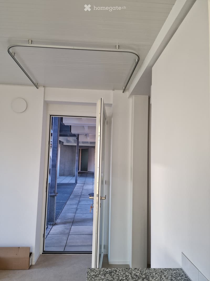 Modern hallway with white walls, a glass door leading to an outdoor area, and a visible staircase in the background.