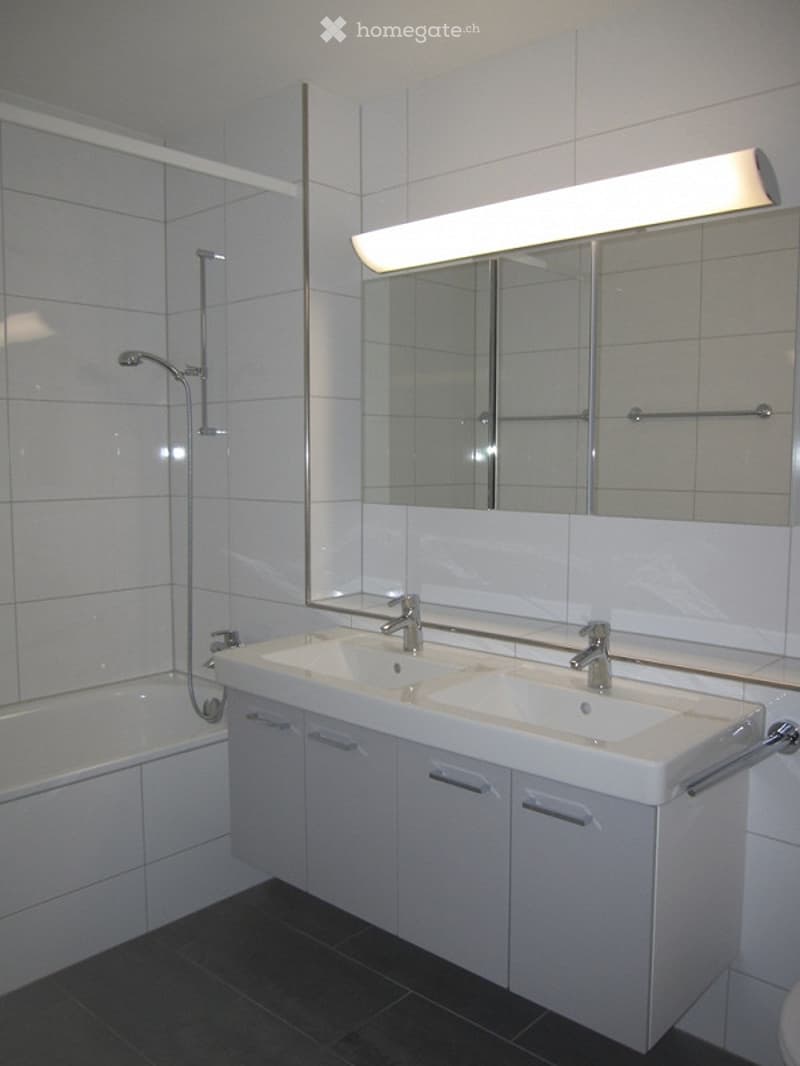 Bathroom with white tiles, bathtub, double sink, mirror, and shower head.