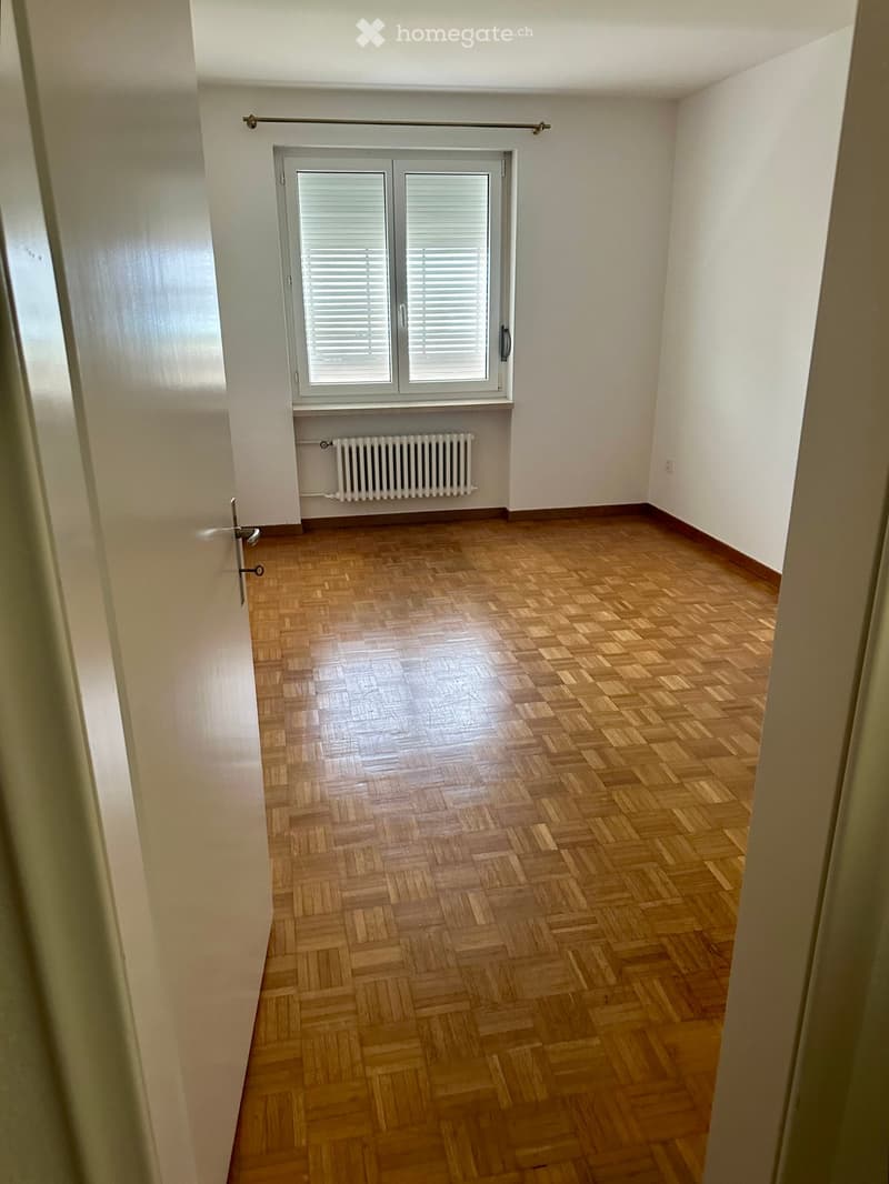 Empty room with wooden parquet flooring, white walls, a window with blinds, and a radiator below it.
