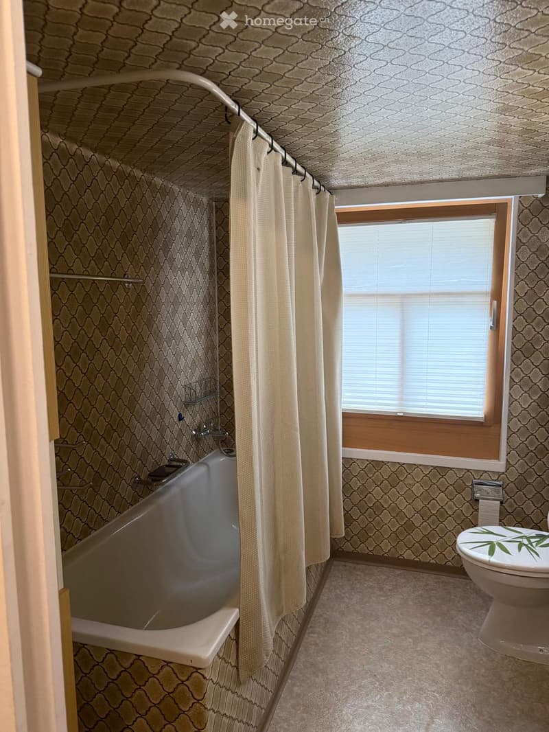 Bathroom with a bathtub, toilet, shower curtain, and window with blinds. The walls and ceiling have a patterned design.