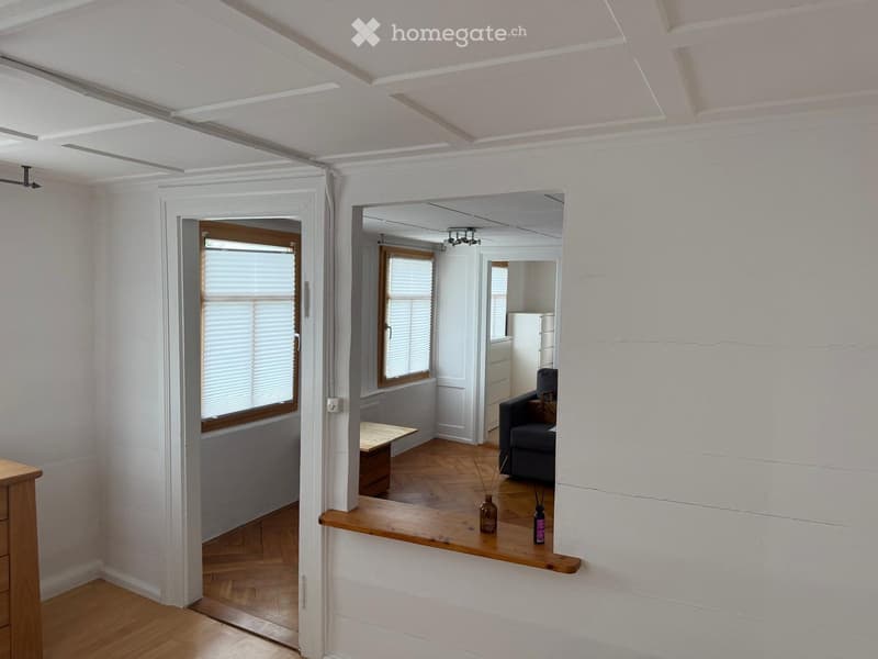 A modern, open-plan living space with wooden flooring, white walls, and large windows. The room features a grey sofa, a small wooden table, and decorative vases on a wooden shelf.