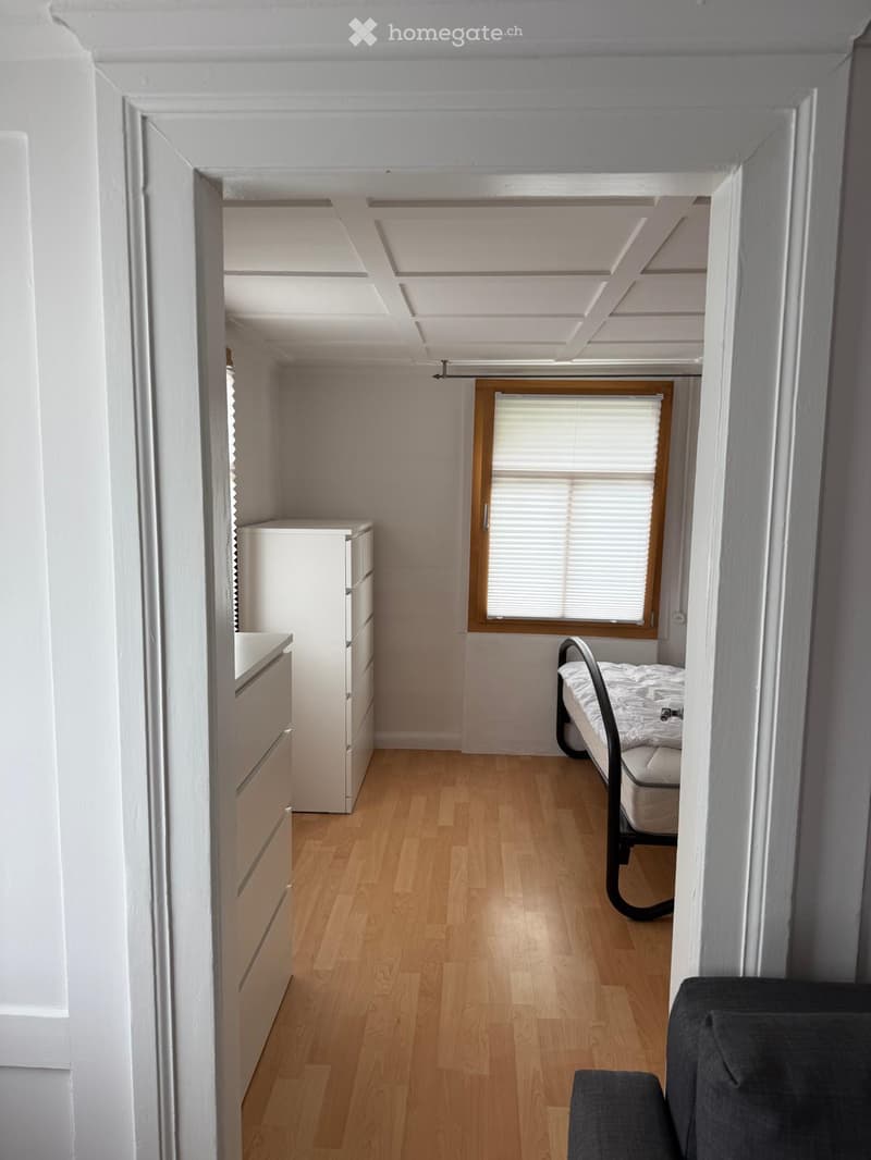 A compact, furnished room with a wooden floor, a bed, a sofa, and a cabinet. The room has white walls and a window with blinds.