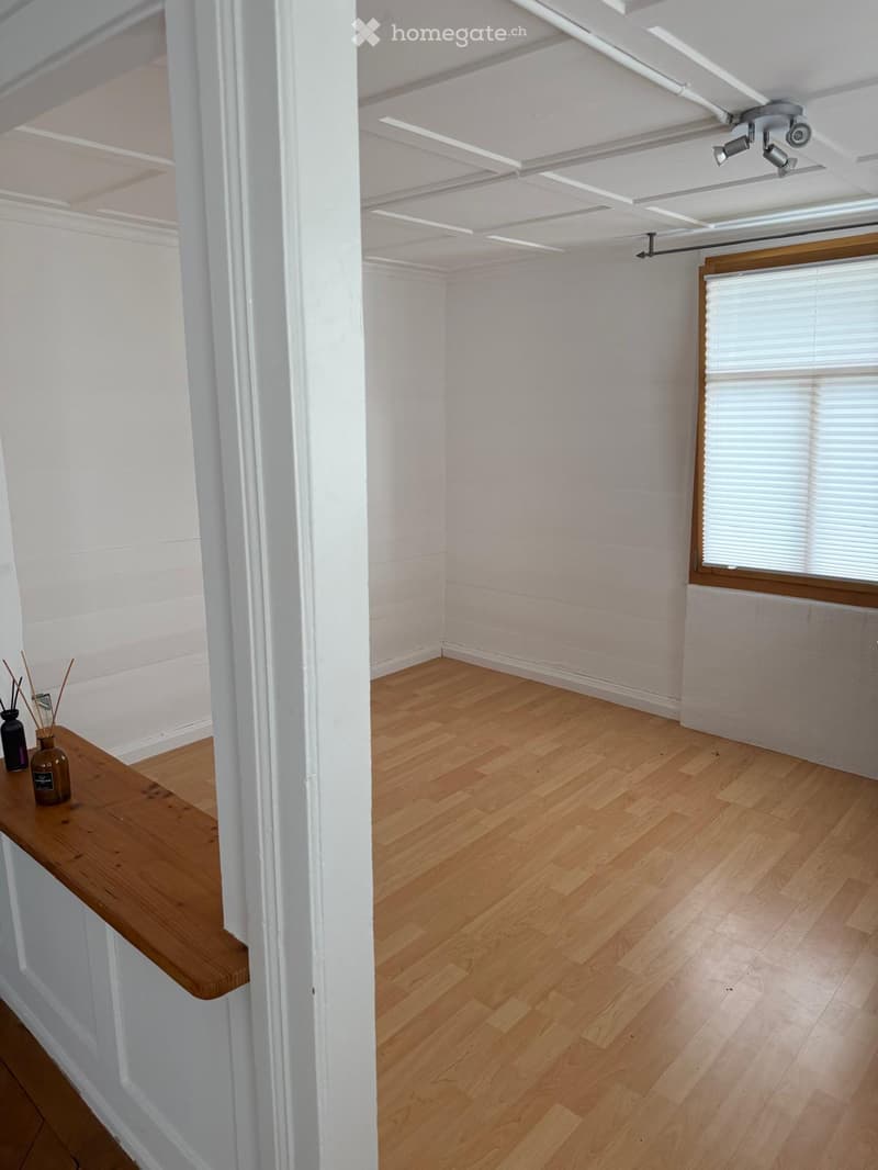 Empty room with wooden flooring, white walls, and a small wooden table with vases on the left side.