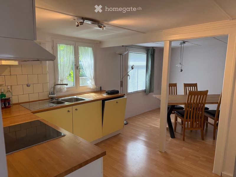The image shows a modern kitchen and dining area with light yellow cabinets, wooden countertops, a sink with a faucet, and a dining table with chairs. The space is well-lit with natural light from windows covered with white curtains.