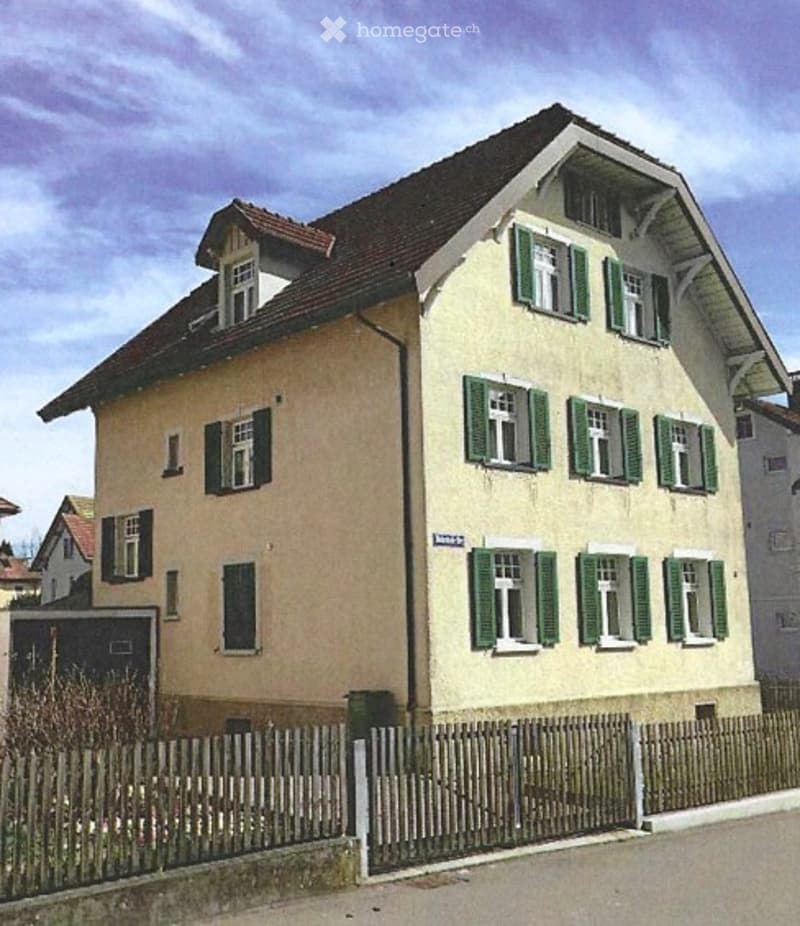 Two-story residential house with beige walls, green shutters, and a dark roof. The house features multiple windows and a small fenced yard in front.