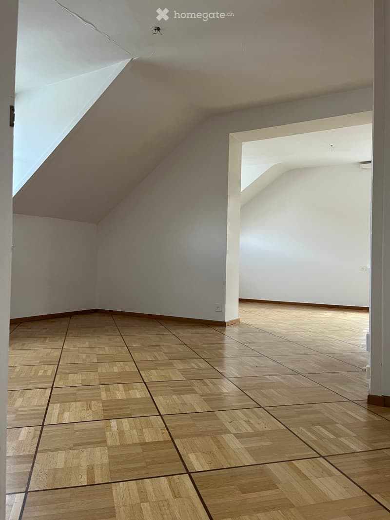 Empty room with wooden parquet flooring and white walls, featuring a slanted ceiling and visible electrical outlets.