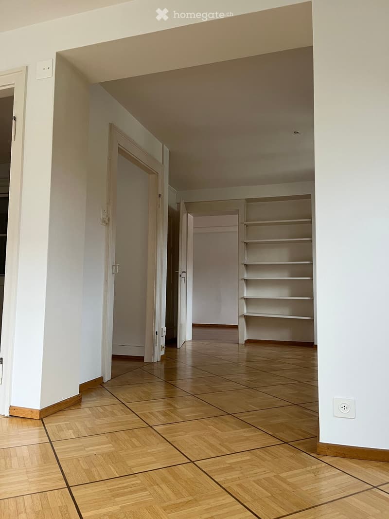 Empty room with wooden flooring, white walls, and open doors leading to other rooms. Shelves are visible in the adjacent room.