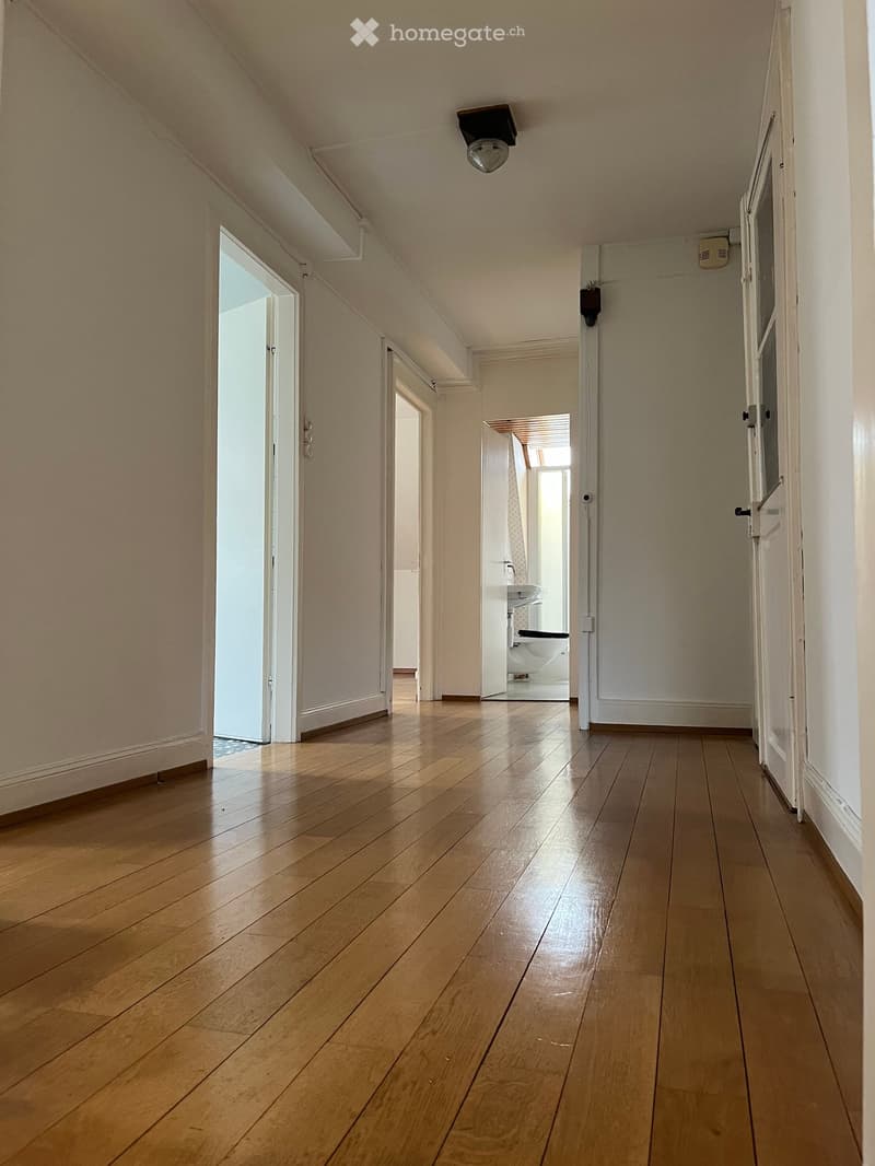 A narrow hallway with wooden flooring, white walls, and several doors leading to different rooms, including a visible bathroom with a sink and toilet.