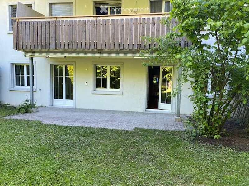 Two-story building with a balcony, white walls, large windows, and a small garden area.
